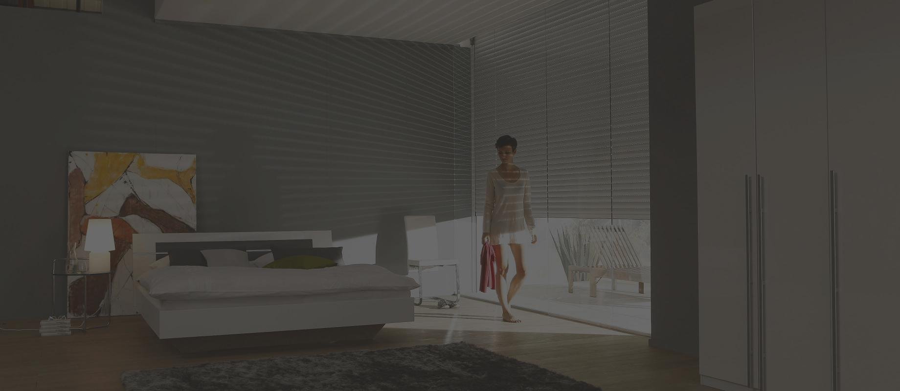 Femme est debout devant la fenêtre de la chambre à coucher, volets roulants à moitié descendus