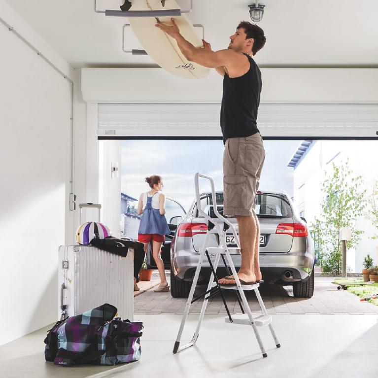Utilisation optimale du garage Homme sur une échelle dans un garage ouvert en train de chercher une planche de surf au niveau du plafond