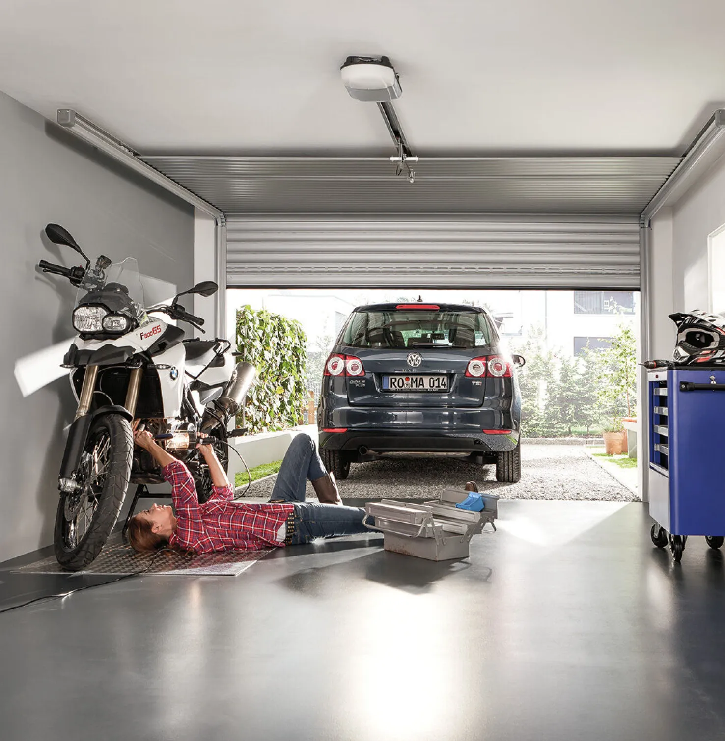 Une femme visse une moto en position couchée dans un garage, une voiture est garée devant.